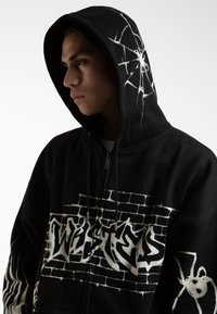 Mustat vetoketjulliset hupparit, joissa on valkoinen graffitotyylinen "TWISTED" -design, seinäkuvio sekä hämähäkkigrafiikka hihassa. Pehmeä kangas.