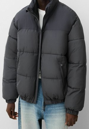 Homme portant une doudoune noire avec une fermeture éclair frontale et des poches latérales zippées sur une chemise blanche et un jean bleu.