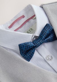 Chemise blanche en tissu texturé, nœud papillon bleu à motifs et col de blazer gris clair texturé visible.