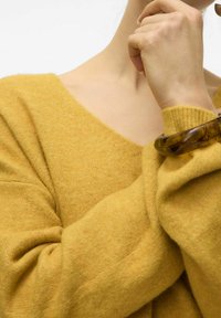 Žlutý pletený svetr s výstřihem do V, který se vyznačuje měkkou texturou a vroubkovanými manžetami. Na zápěstí je nošena hnědá kulatá ozdoba.