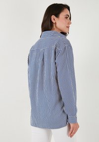 Camicetta a righe blu e bianche con maniche lunghe, colletto con bottoni, vestibilità rilassata e dettaglio a piega sul retro. Tessuto liscio, polsini su misura e orlo.