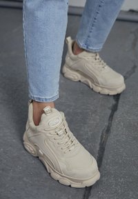 Beige-tennis kengät, joissa on teksturoitu pinta, nauhakiinnitys ja pehmustettu nilkatuki. Paksu kumipohja, jossa on brändätyt yksityiskohdat ja logo.