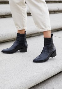 Botas negras hasta el tobillo con textura de croc embossing, puntas afiladas y un tacón bajo en bloque. Combinadas con pantalones crema arremangados en los escalones.