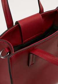 Punainen nahkalaukku, jossa on sileä pinta, metallinen rengas yksityiskohtana ja musta sisus. Vankat kahvat ja selkeät linjat.