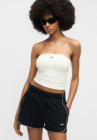 Frau mit großem Afro, die schwarze Sonnenbrille, ein weißes trägerloses Nike-Oberteil und schwarze Nike-Shorts mit weißem Rand trägt, steht mit einer Hand in der Tasche.