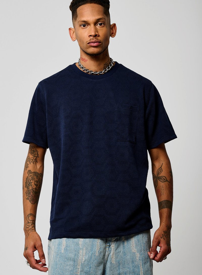 T-shirt bleu marine à manches courtes avec un motif hexagonal texturé, col rond et poche poitrine unique. Porta avec un short en denim clair déchiré.