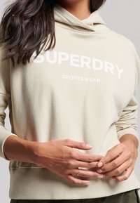Beige mikina s kapucí s potiskem "SUPERDRY" a "SPORTSWEAR" v bílé barvě. Měkký materiál s volným střihem a žebrovanými manžetami.