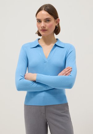 Femme aux cheveux bruns courts portant un pull bleu clair à col et un pantalon gris à fines rayures, les bras croisés, fond neutre.