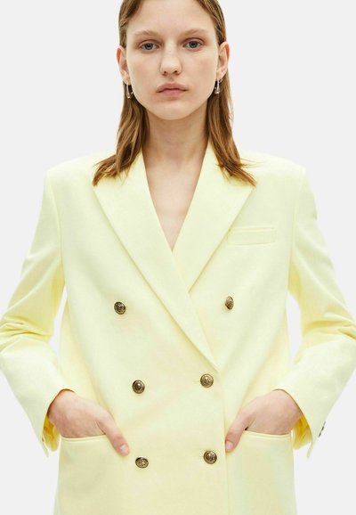 Dubbelzijdige blazer van lichtgele stof, met gouden knopen, een enkele borstzak en klepzakken aan beide zijkanten. Soepele textuur.