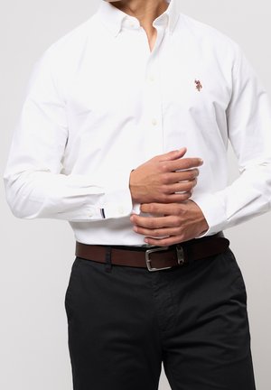 Mężczyzna poprawiający mankiet białej koszuli zapinanej na guziki z wyhaftowanym logo, ubrany w czarne spodnie i brązowy pasek.