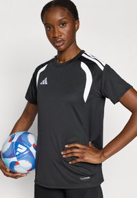 Μαύρο αθλητικό μπλουζάκι με λευκές λεπτομέρειες, στρογγυλή λαιμόκοψη και ύφασμα που απορροφά την υγρασία. Στο χέρι κρατιέται μια μπλε και λευκή μπάλα ποδοσφαίρου.