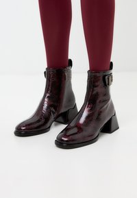Bottines bordeaux brillantes avec un talon bloc géométrique, fermeture éclair et accent de boucle. Présente un aspect cuir lisse et texturé.