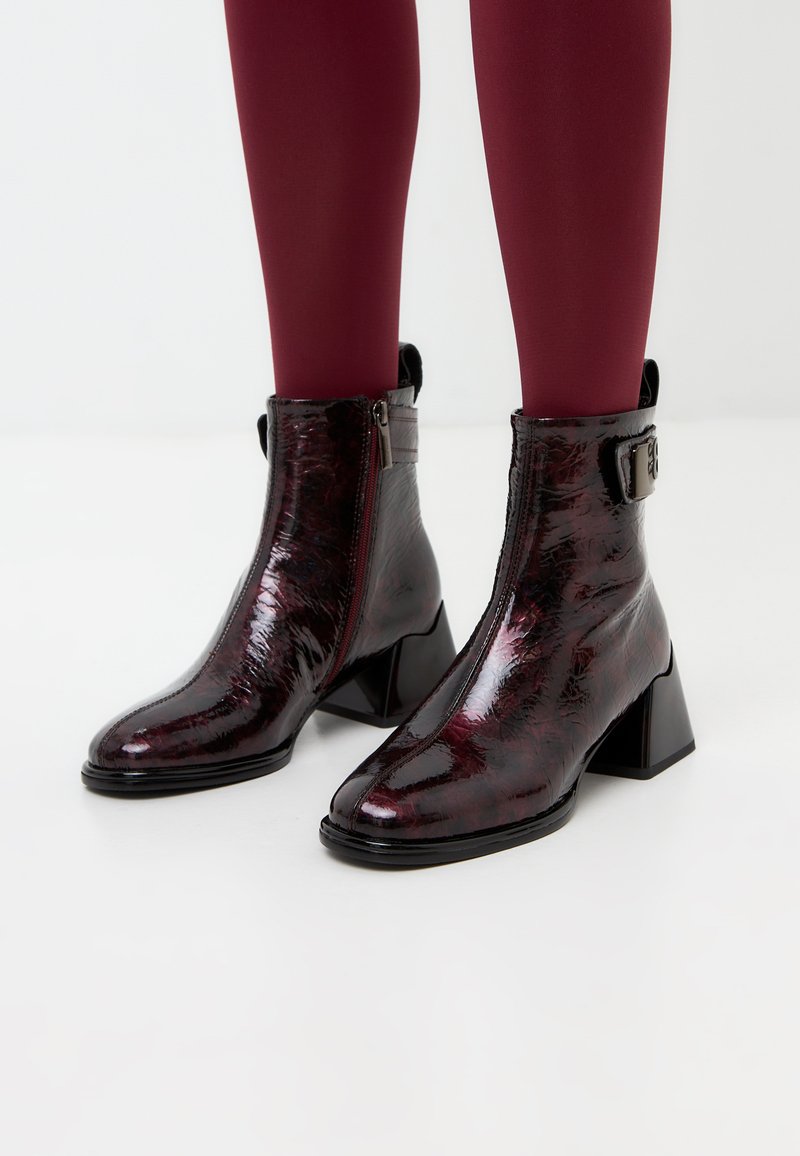 Bottines bordeaux brillantes avec un talon bloc géométrique, fermeture éclair et accent de boucle. Présente un aspect cuir lisse et texturé.