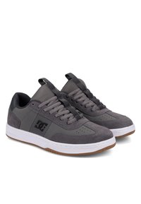 Pari matalakoristeisia harmaita lenkkareita mustalla logolla, verkko- ja mokkanahkakomponenteilla, mustilla nauhoilla, valkoisilla pohjilla ja ruskealla kumipohjalla valkoista taustaa vasten.