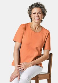 T-shirt met korte mouwen en ronde hals van levendig oranje katoen, met een gladde textuur. Model zittend op een houten stoel, gekleed in witte broek.