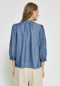 Blusa blu denim con maniche a sbuffo 3/4, scollatura arricciata, texture liscia e un leggero lucido. Presenta un orlo dritto e polsini con bottoni.