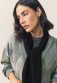 Grå sweater med grønne diamantmønstre, kombineret med et sort halstørklæde. Glat tekstur, afslappet design, let oversized pasform.