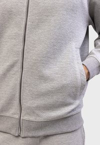 Siva zip-up dukserica s teksturiranim materijalom, bočnim džepovima, rebrastim manžetama i donjim rubom, te glatkim patentnim zatvaračem sprijeda.