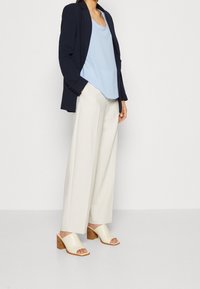 Granatowa marynarka na jasnoniebieskiej bluzce bez rękawów, w zestawieniu z białymi spodniami szerokimi i beżowymi klapkami na słupku z drewnianymi akcentami.