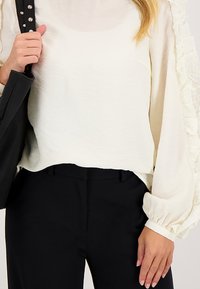 Witte textuur blouse met volumineuze mouwen, gecombineerd met zwarte op maat gemaakte broeken. Zwarte handtas met genagelde band.