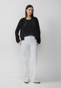 Zwarte geplooide bomberjack over een zwart top, gecombineerd met witte wijde broek en beige sneakers; gladde texturen en gestroomlijnde vormen.