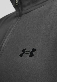 Graue Zip-Jacke aus glattem Stoff, mit einem schwarzen Under Armour-Logo auf der linken Seite. Nahaufnahme von Textur und Details.