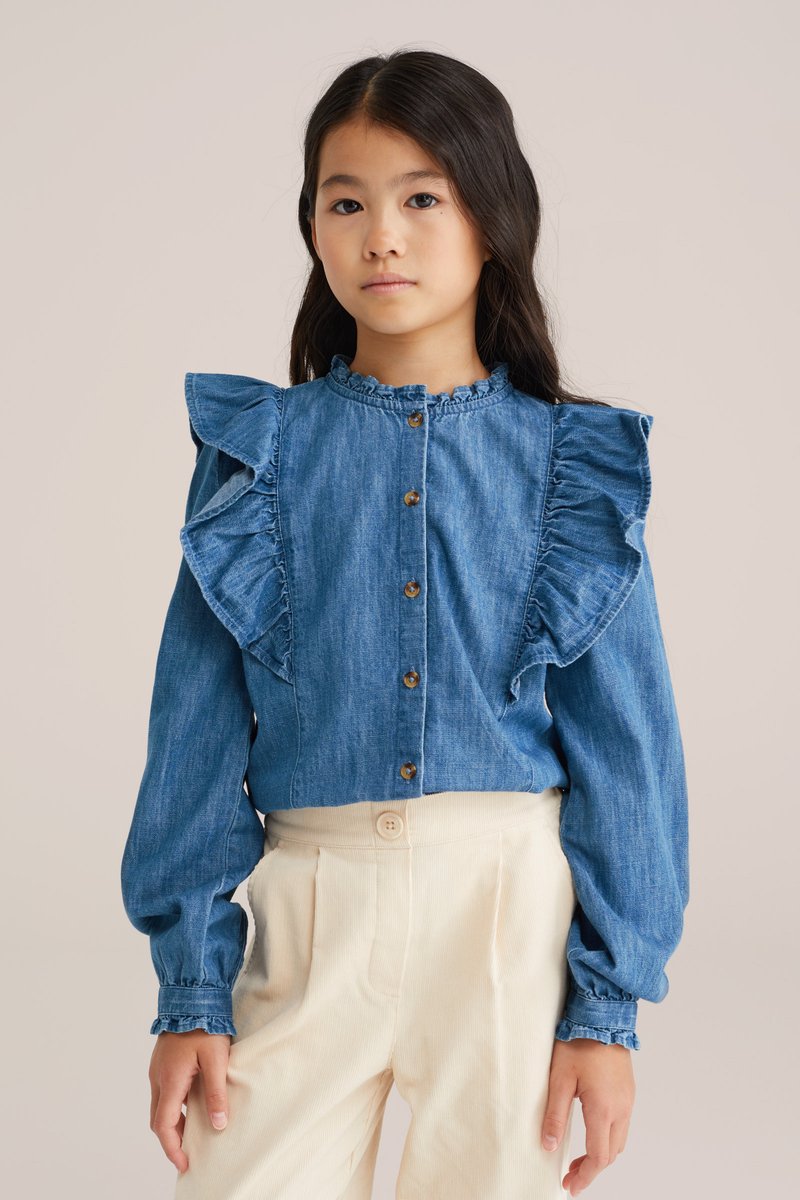 Denim bluza z nabranimi rokavi, gumbom za zapiranje in okroglim izrezom. Ima svetlo modro pranje in mehak občutek, v kombinaciji s smetanastimi hlačami.