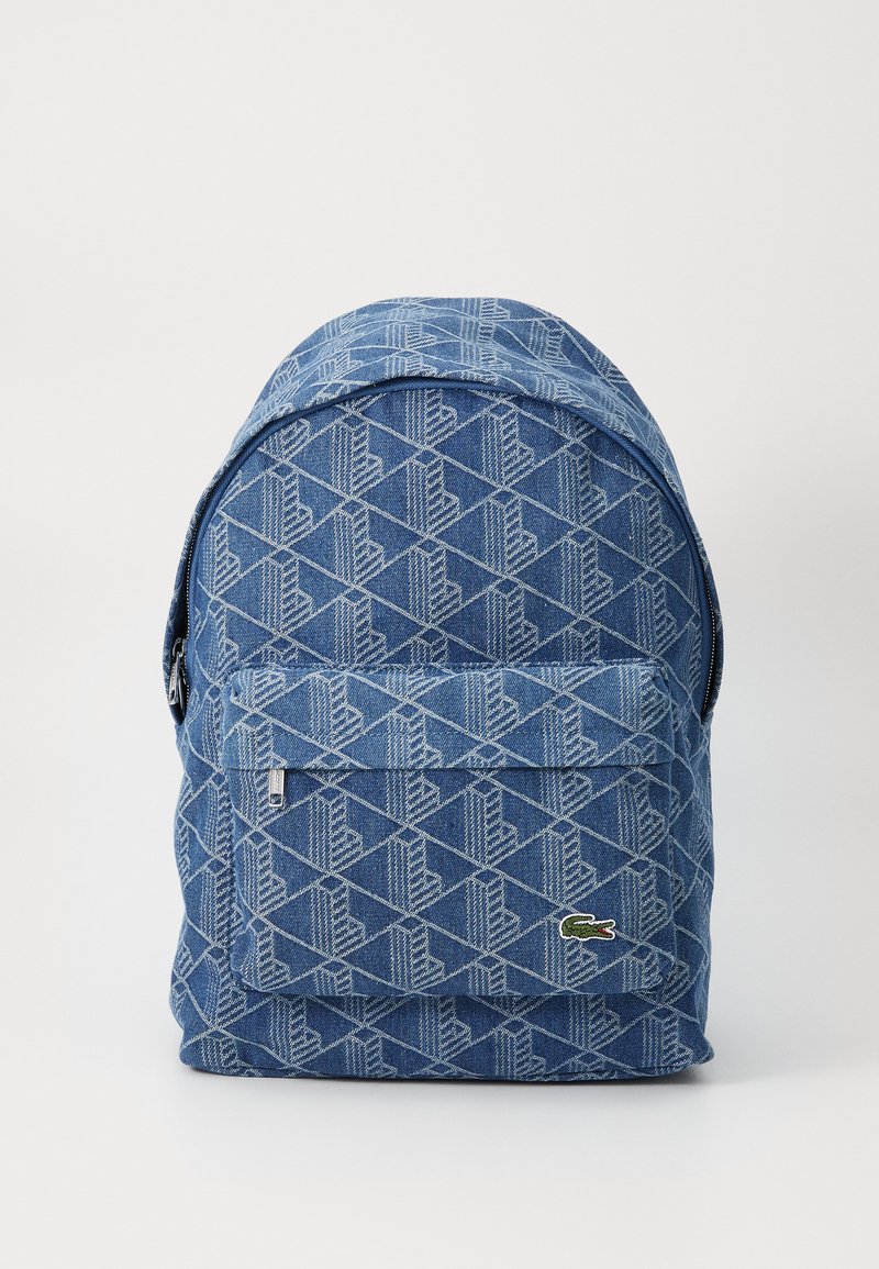 Mochila de denim com padrão geométrico em azul claro e azul escuro, com um bolso frontal com fecho e um pequeno patch com logotipo na parte inferior.