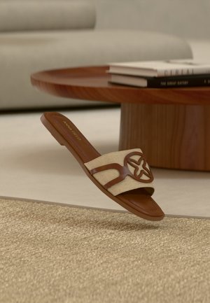 Sandale à glissière marron et beige avec une sangle tissée présentant un motif à découpe circulaire, placée près d'une table basse ronde en bois avec des livres empilés.