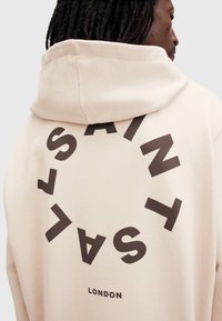 Hanorac cu glugă de culoare bej deschis, cu un imprimeu circular mare care conține „SAINT” și „LONDON” în brun închis. Material moale și design casual.