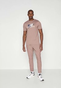 Lys rosa treningst-skjorte med marineblå "New Balance" logo, kombinert med matchende tapered bukser og hvite sneakers. Enkel, glatt stoffdesign.