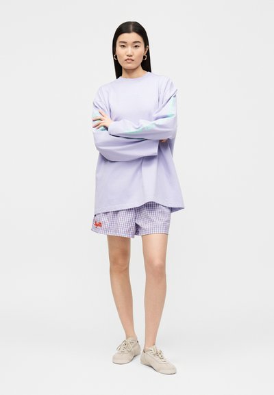 Νεαρή γυναίκα στέκεται με σταυρωμένα τα χέρια, φοράει φαρδιά μωβ φούτερ, καρό σορτς, μπεζ αθλητικά παπούτσια και χρυσούς κρίκους στα αυτιά.