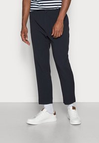 Granatowe spodnie wizytowe o dopasowanym kroju, z zakładkami z przodu i zwężanymi nogawkami, zestawione z białymi tenisówkami i jasnoszarymi skarpetkami.