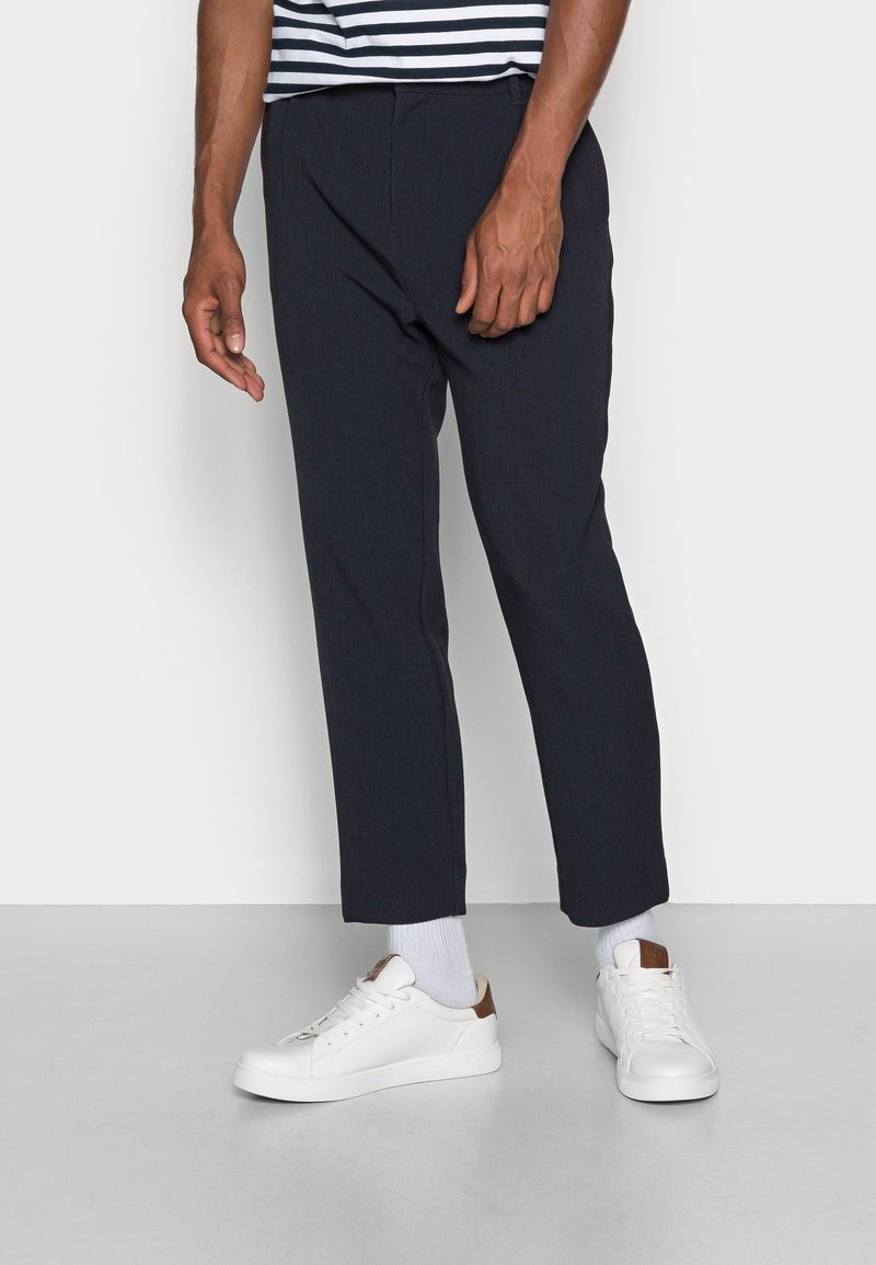 Granatowe spodnie wizytowe o dopasowanym kroju, z zakładkami z przodu i zwężanymi nogawkami, zestawione z białymi tenisówkami i jasnoszarymi skarpetkami.