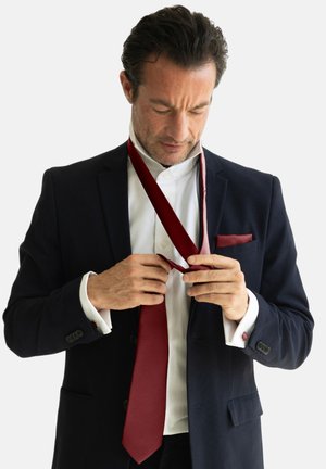 Un homme en blazer bleu marine et chemise blanche ajuste une cravate bordeaux. Le costume a une texture lisse avec des boutons détaillés et un mouchoir de poche.