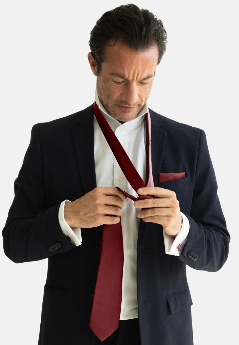 Un hombre en una chaqueta azul marino y camisa blanca ajusta una corbata burdeos. El traje tiene una textura suave con botones detallados y un pañuelo de bolsillo.