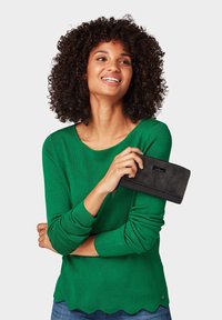 Maglia verde a maniche lunghe lavorata a maglia con orlo scallop, abbinata a un portafoglio in pelle nera con chiusura a zip e cuciture a contrasto.