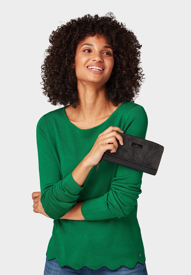 Maglia verde a maniche lunghe lavorata a maglia con orlo scallop, abbinata a un portafoglio in pelle nera con chiusura a zip e cuciture a contrasto.