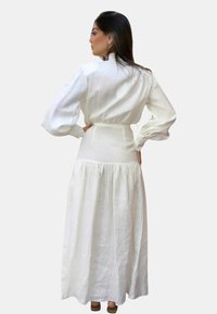 Abito maxi in lino bianco con maniche a sbuffo, vita aderente e gonna a strati. Presenta una sottile texture e un dettaglio con fiocco sulla schiena.