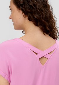 Roze top met korte mouwen, gemaakt van een textuurstof, met een V-halslijn en gekruiste bandjes op de rug, die een eenvoudige maar onderscheidende uitstraling benadrukt.