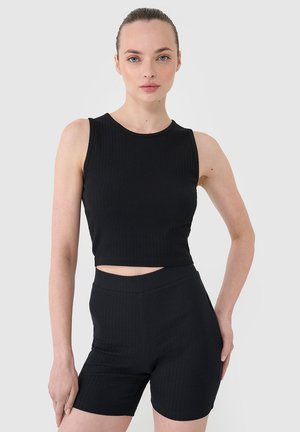 Kobieta z włosami związanymi do tyłu nosi czarny bezrękawnik typu crop top i pasujące wysoko taliowane rowerowe szorty, stoi na tle jednolitego tła.