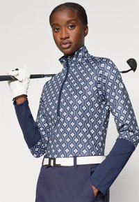 Tengerészeti mintás golf pulóver magas gallérral és negyed cipzárral. Geometrikus fehér mintákkal és kontrasztos tengerészkék ujjal rendelkezik. Bézs övvel viselve.