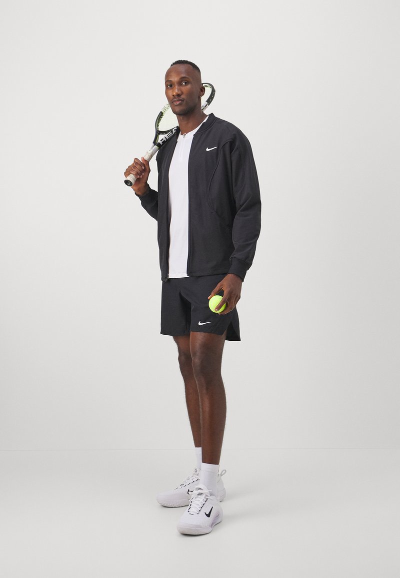 Zwarte Nike-jack met wit logo, witte t-shirt, zwarte shorts, met een tennisracket en een groene tennisbal, staand op een effen achtergrond.