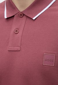 Gros plan d'une personne portant un polo rose avec deux boutons et une bande blanche sur le col, avec un écusson logo "BOSS".