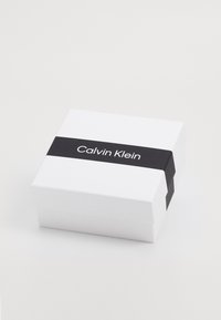 Baltas stačiakampis dėžutė su juoda juosta, ant kurios užrašyta „Calvin Klein“. Lygus paviršius, minimalistiškas dizainas, matinė apdaila.