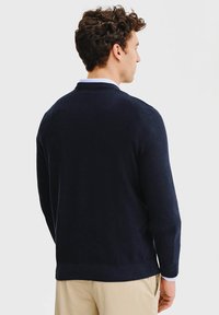 Granatowy sweter z teksturowanego dżerseju z prążkowanym kołnierzem i mankietami, o gładkim wzorze i dopasowanej sylwetce, noszony na jasnej koszuli.