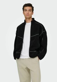 Veste en polaire noire avec une fermeture éclair à l'avant, un col arrondi et des coutures grises contrastantes. Assortie à un pantalon beige.