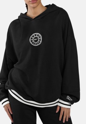 Femme portant un sweat à capuche Reebok noir ample avec un logo blanc sur la poitrine et des poignets rayés, associé à un pantalon noir.
