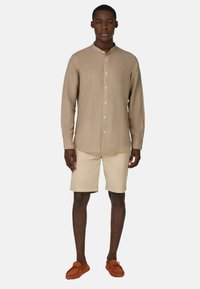 Camisa de manga larga color marrón claro con cuello mao y cierre de botones en el frente, combinada con pantalones cortos beige hasta la rodilla y zapatos de estilo mocasín marrones.