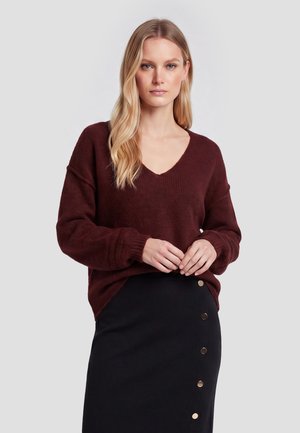Lyshåret kvinde med løst hår iført en mørkerød V-hals sweater og et sort skørt med metal knapper foran.